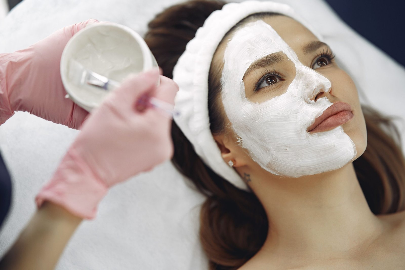 From above closeup of crop faceless cosmetologist applying white clay mask with brush to female clients face during procedure in spa center